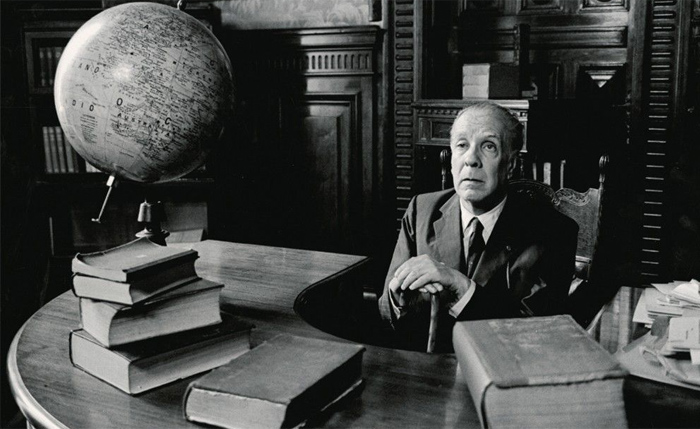Jorge Luis Borges sentado ante un escritorio semicircular sobre el que hay un globo terráqueo, ambas manos de Borges reposan sobre su batón, colocado frente a él